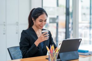 Woman in blazer at table looking at iPad while drinking coffee