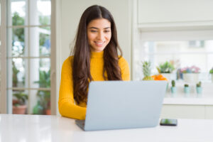 A woman wearing a yellow turtle smiling at a laptop